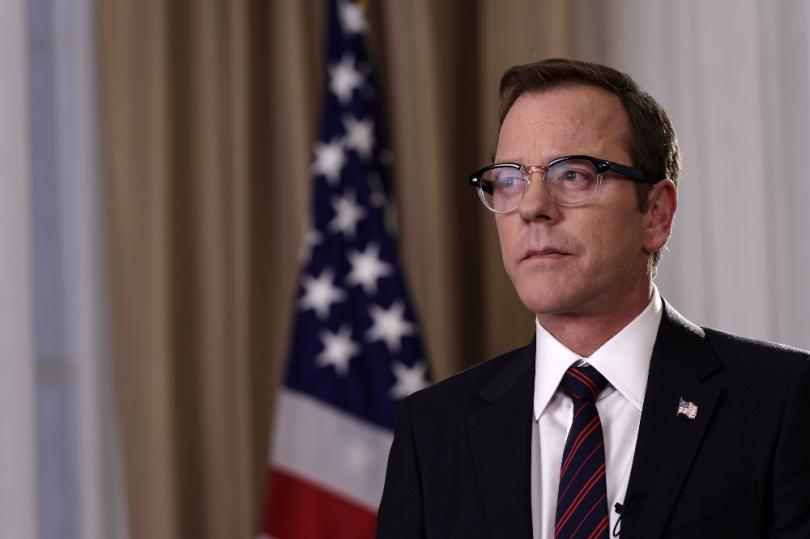 Man in suit and glasses in front of flag.