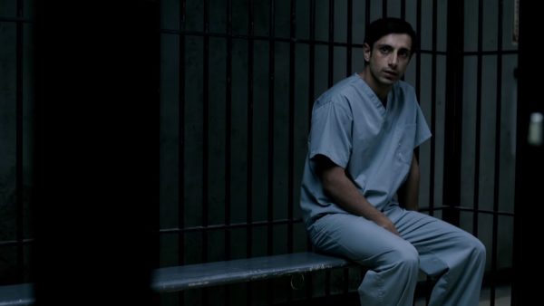 Man in blue jumpsuit sits in jail cell.