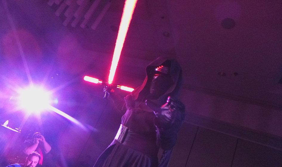 Person in hooded costume holding lightsaber.