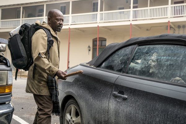 Man with a stick near a car with a zombie inside.