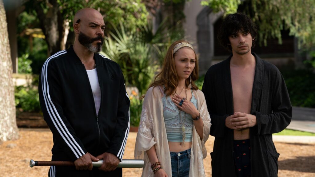 A man with a baseball bat in a track suit, a girl in a crop top, and a shirtless man with a bathrobe stare into the distance