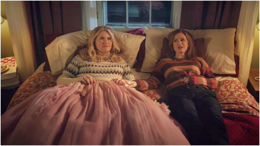 Jillian Bell and Isla Fisher relax on a bed in the film GODMOTHERED on Disney+