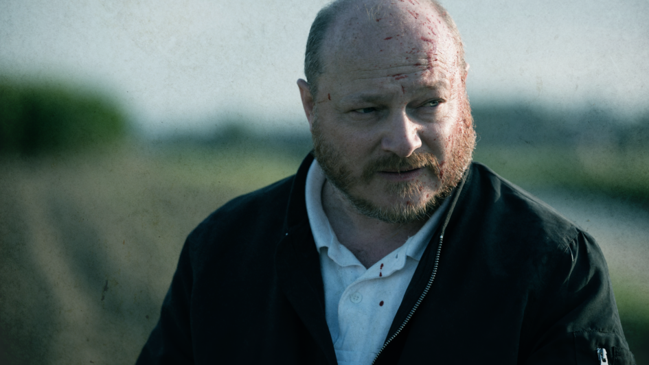 Olivier Picard as Scola, his face bloodied, wearing a black jacket and white shirt in the psychological horror film Heat of Madness (2026).