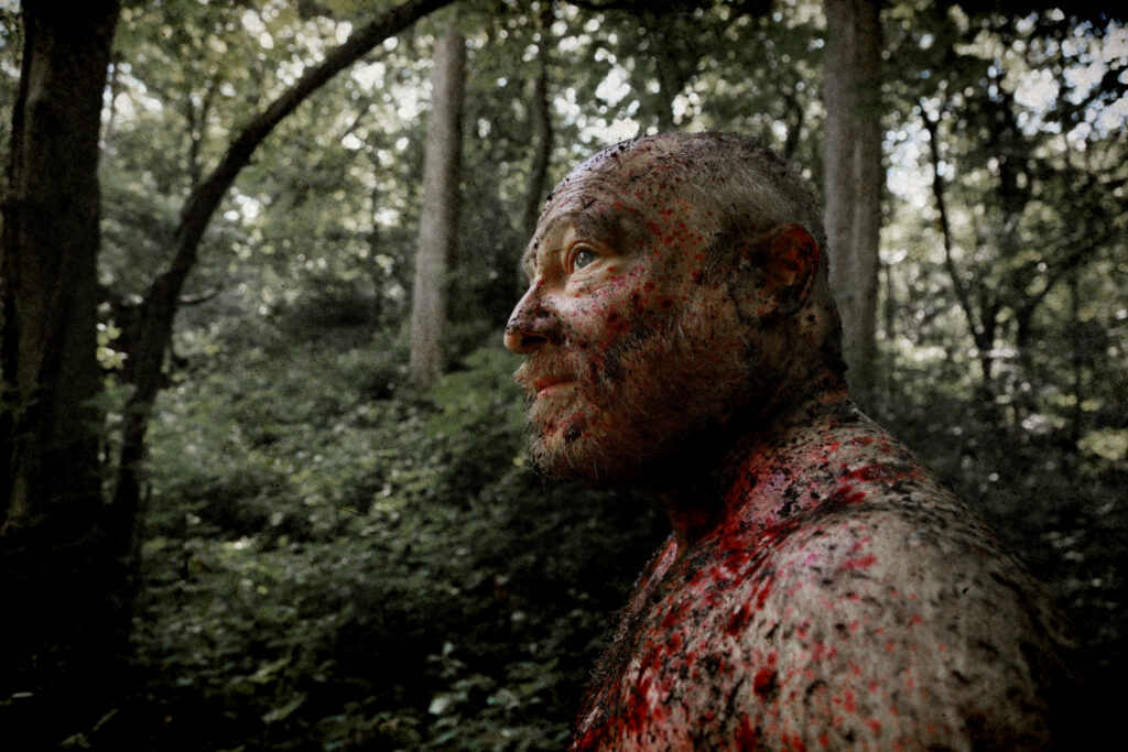 Blood-covered Scola, portrayed by Olivier Picard, looks upward while standing in a wooded area in Heat of Madness (2026).